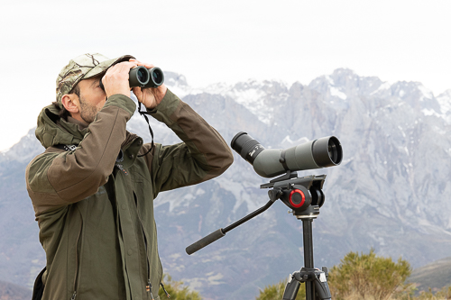 Observador con prismáticos junto a un telescopio, con montañas nevadas al fondo